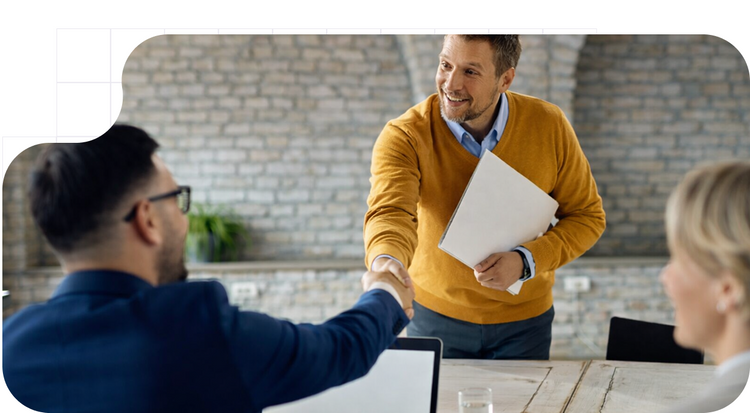 Hombre de pie con suéter mostaza estrechando la mano de un hombre con traje azul sentado frente a un portátil tras firmar un acuerdo de confidencialidad